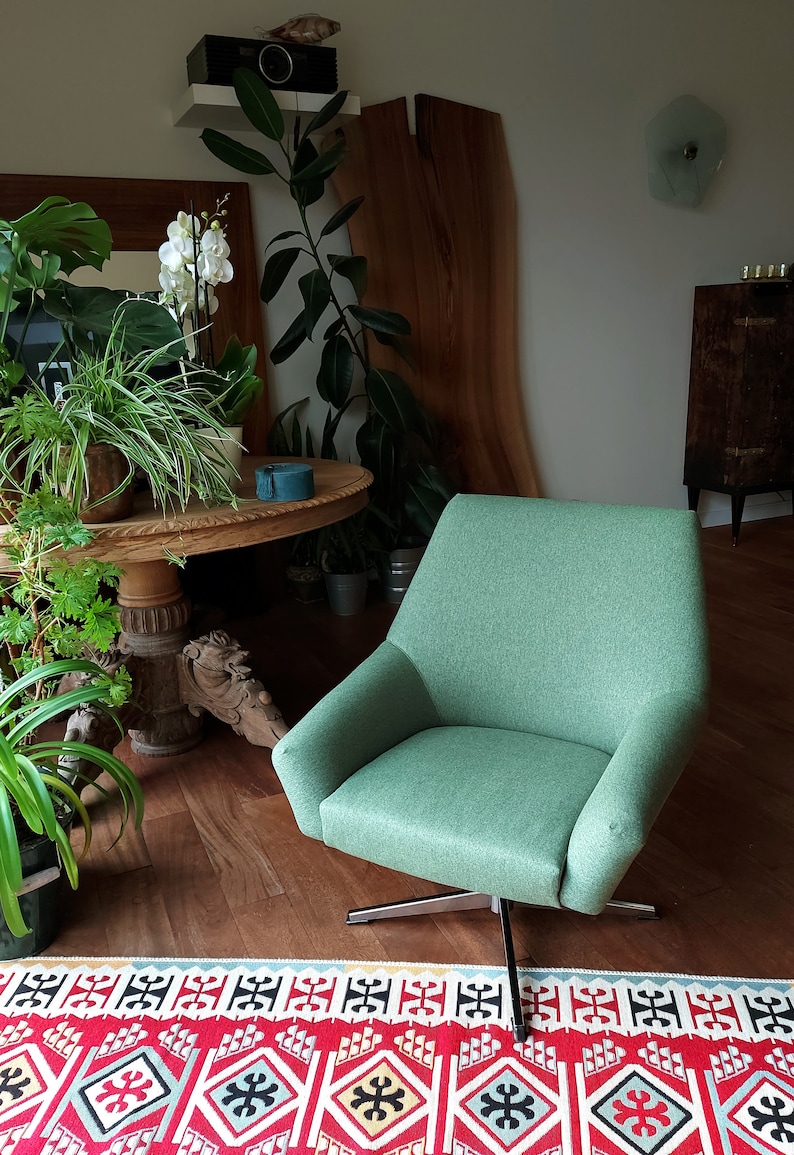 May include: A green swivel chair with a modern design sits on a red and white patterned rug in a living room. The chair is in front of a wooden table with plants and a wooden wall panel.