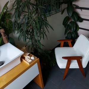 May include: A white armchair with wooden arms and legs sits in a room with plants and a white sink. The armchair has a textured fabric and the wooden arms and legs are a dark brown color.