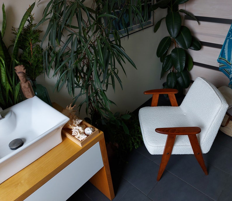 May include: A white armchair with wooden arms and legs sits in a room with plants and a white sink. The armchair has a textured fabric and the wooden arms and legs are a dark brown color.