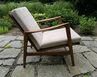May include: A wooden armchair with a light gray fabric cushion. The chair is sitting on a stone patio.