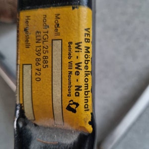 May include: A yellow label with black text on a black metal bar. The label reads "VEB M&ouml;belkombinat Wi-We-Na Betrieb VIII Naumburg" and "Modell nad: TGL 25 885 ELN 139 86 720 Hergestellt".