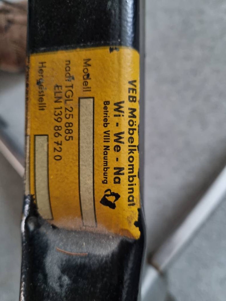 May include: A yellow label with black text on a black metal bar. The label reads "VEB M&ouml;belkombinat Wi-We-Na Betrieb VIII Naumburg" and "Modell nad: TGL 25 885 ELN 139 86 720 Hergestellt".