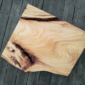 Könnte beinhalten: Eine große, natürliche Holzplatte mit einer lebenden Kante und einem einzigartigen Maserungsmuster. Das Holz ist hellbraun mit dunkleren braunen Streifen und Knoten. Die Platte ist glatt und poliert.