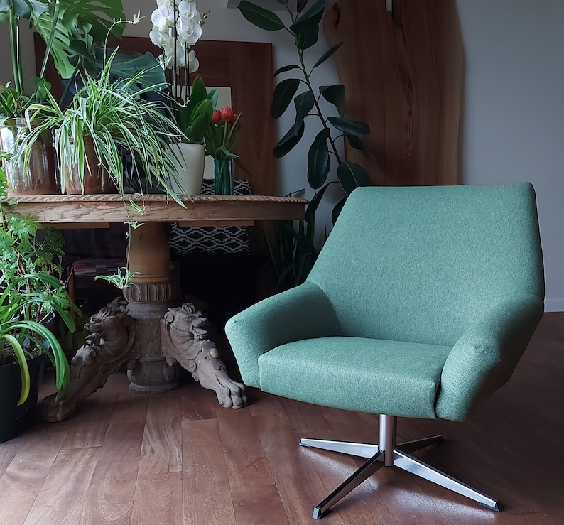 May include: A green swivel chair with a wooden base and a carved design. The chair is in a living room setting with plants and a table in the background.