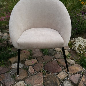 May include: A beige upholstered armchair with a rounded back and black metal legs. The chair is sitting on a stone patio.