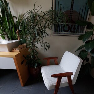 May include: A white armchair with wooden arms and legs sits in a room with plants and a sink. The chair has a plush, white fabric covering.