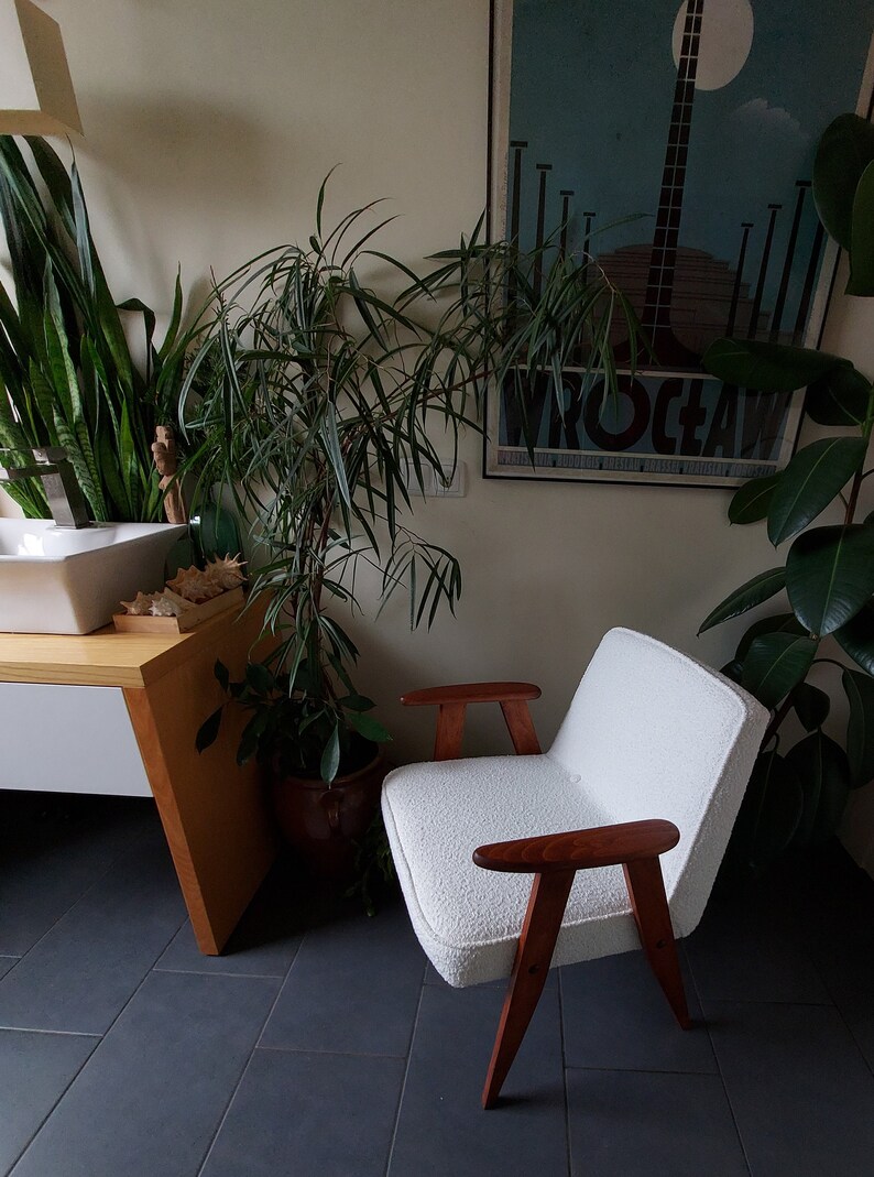 May include: A white armchair with wooden arms and legs sits in a room with plants and a sink. The chair has a plush, white fabric covering.