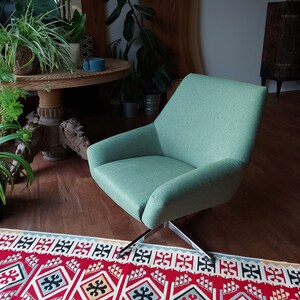 May include: A light green swivel chair with a black metal base sits on a red, white and black patterned rug. The chair is in a living room setting with plants and a wooden table in the background.