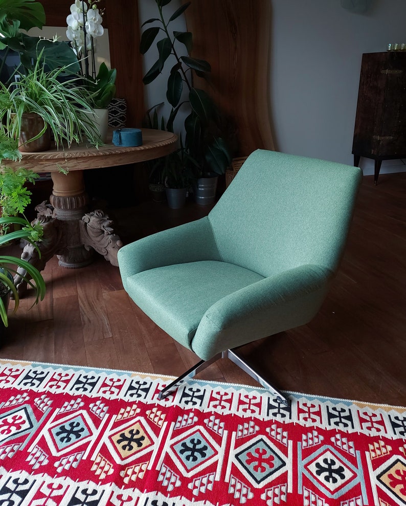 May include: A light green swivel chair with a black metal base sits on a red, white and black patterned rug. The chair is in a living room setting with plants and a wooden table in the background.