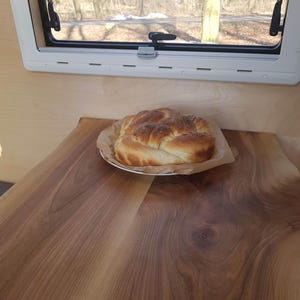 Könnte beinhalten: Ein frisch gebackenes Brot liegt auf einem Holztisch vor einem Fenster mit Blick auf einen Wald. Das Brot ist goldbraun und hat ein geflochtenes Muster.