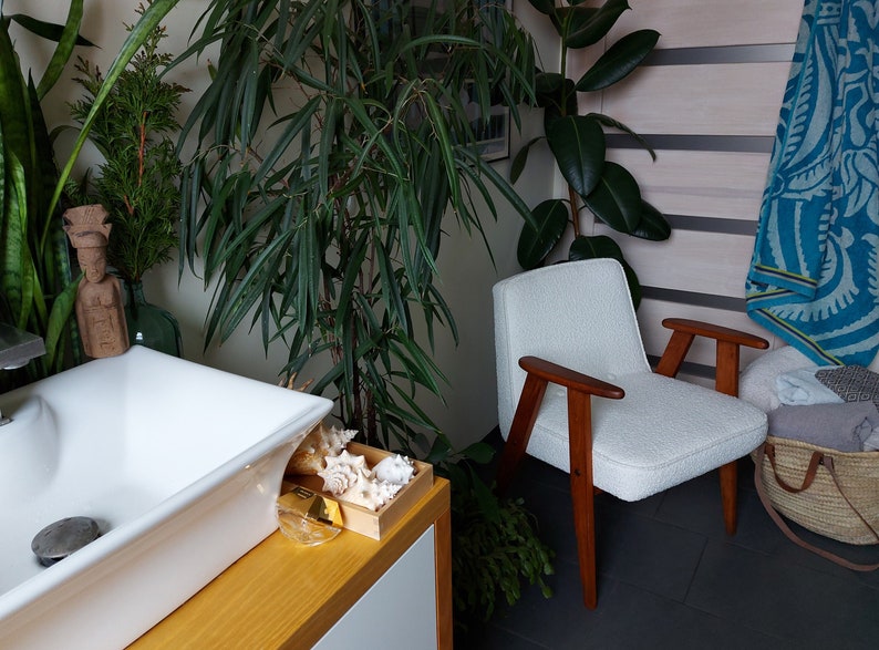 May include: A white armchair with wooden arms and legs sits in a bathroom with a white porcelain sink and a wooden countertop.  The bathroom has a variety of plants, including a large green plant in the corner and a smaller plant on the countertop.  There is a blue and white patterned towel hanging on the wall.