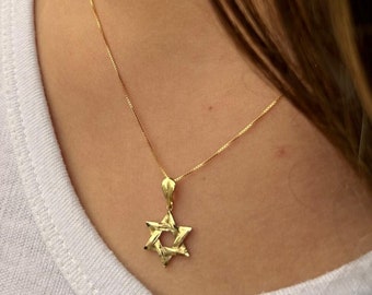 May include: A gold-coloured Star of David pendant necklace. The pendant is a six-pointed star with a textured surface, suspended from a delicate gold chain. The necklace is worn against a white shirt.