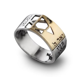 May include: A silver ring with a gold-colored Star of David design. The ring has Hebrew text engraved on the band. The ring is a close-up shot on a white background.
