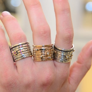 Puede incluir: Una mano con cuatro anillos de plata con texto grabado y pequeñas piedras preciosas. Los anillos están apilados en cada dedo.