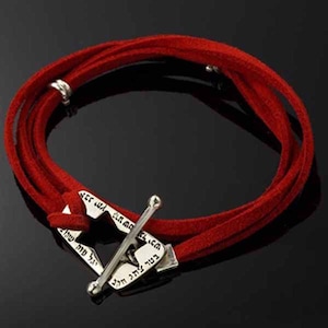 May include: Red suede wrap bracelet with a silver-colored clasp. The bracelet wraps around multiple times and features Hebrew text on the clasp. The clasp has a bar that secures the bracelet.