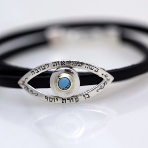 May include: A silver Hamsa hand charm with a turquoise stone in the center, set on a black leather bracelet. The charm is inscribed with Hebrew text.