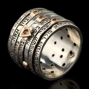 May include: Silver-toned spinner ring with Hebrew inscriptions. The ring features multiple bands, some with etched text, and is accented with gold-colored details and small red gemstones. The design includes a triangular gold accent with a red stone.