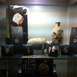 May include: A display case with various silver necklaces and pendants, including a pendant with a tree of life design. There are also two ceramic vases, one with a brown and white glaze and the other with a brown glaze. The display case is lit with overhead lights. The text "a Gift of Love Hope & Prayer" is visible on a box in the lower right corner.