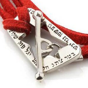 May include: A silver bracelet with a red suede band. The bracelet features a geometric design with Hebrew text. The clasp is a T-bar style. The bracelet is shown against a white background.