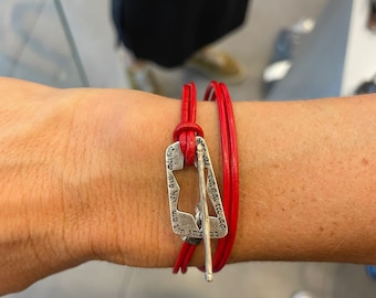 May include: Red leather bracelet with a silver clasp. The bracelet wraps around the wrist twice and features a rectangular silver clasp with engraved text. The clasp has a bar that goes through a loop to secure the bracelet.