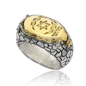 May include: A silver ring with a textured band and a gold oval top. The top features a Star of David and the Hebrew word "שלום" (peace) in gold.