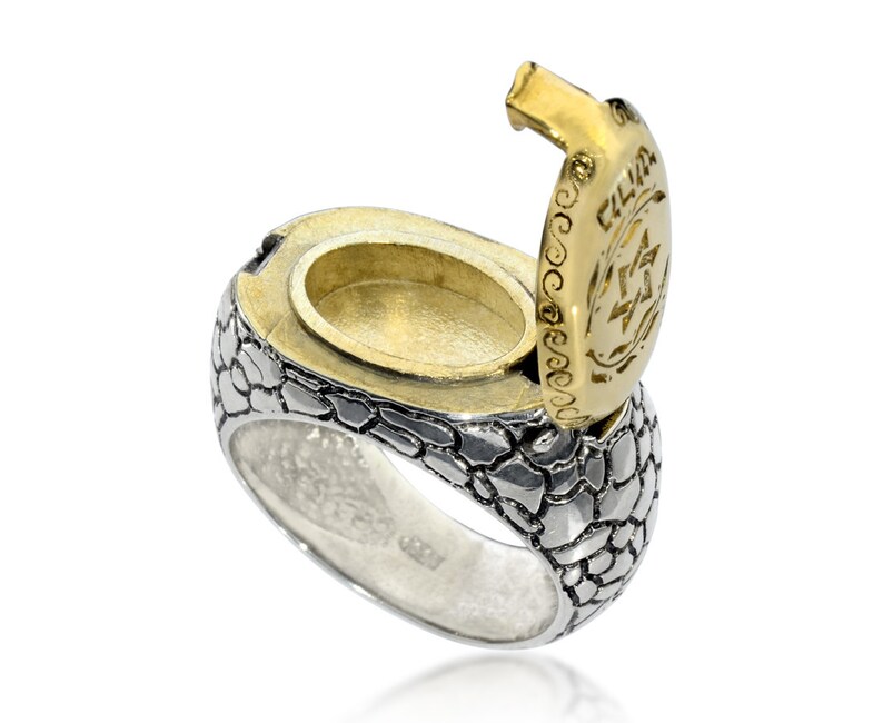 May include: A silver ring with a textured, scaly design. The ring has a hinged, gold-plated top that opens to reveal a small compartment. The top is engraved with a Star of David and the Hebrew word "Chai".