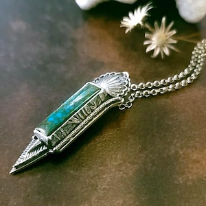 May include: A silver pendant necklace featuring a rectangular turquoise stone. The pendant has intricate metalwork with a pointed bottom and a shell-like design at the top. The necklace has a delicate chain.