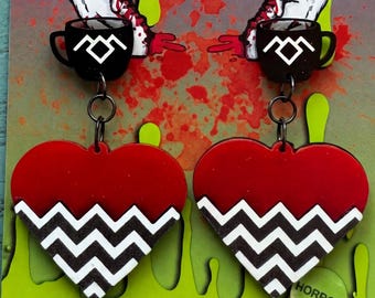 May include: A pair of dangle earrings featuring a red heart shape with a white zigzag design. The earrings are attached to a black cup with a white symbol. The background has the text "SEAGRIFFIN CREATIONS" and the word "HORROR".