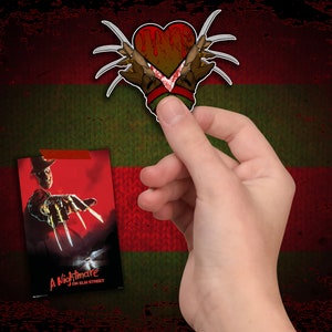 May include: A red heart with a bloody design and Freddy Krueger's gloved hand with blades coming out of it. The heart is on a green and red striped background. The text "A Nightmare on Elm Street" is on a poster in the background.