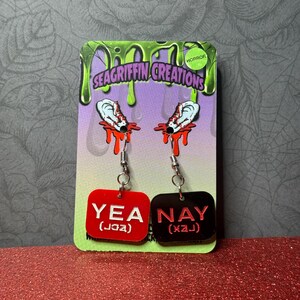 A pair of horror-themed earrings displayed on a green and purple card. Each earring features a white ear with red blood dripping, and a red or black rectangular charm with the words 'YEA' or 'NAY' in white. The card has 'SEAGRIFFIN CREATIONS' and 'HORROR' printed on it. My Chemical Romance, The Black Parade, MCR, Gerard Way