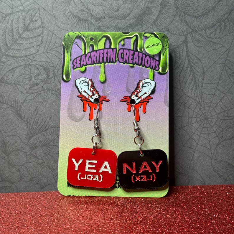 A pair of horror-themed earrings displayed on a green and purple card. Each earring features a white ear with red blood dripping, and a red or black rectangular charm with the words 'YEA' or 'NAY' in white. The card has 'SEAGRIFFIN CREATIONS' and 'HORROR' printed on it. My Chemical Romance, The Black Parade, MCR, Gerard Way