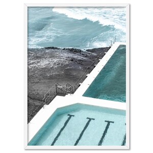 May include: An aerial view of a swimming pool with three lanes, situated next to a rocky ocean shore with crashing waves. The pool is surrounded by white concrete and has a metal ladder leading up to the deck.