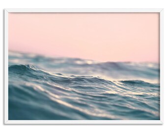 May include: A framed print featuring a close-up view of ocean waves. The water is a mix of blue and teal, with gentle ripples and a soft, blurred pink sky in the background. The white frame adds a clean, modern touch.