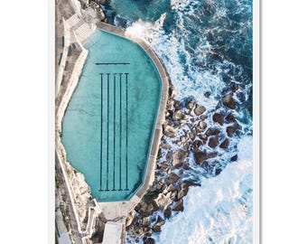 以下が含まれることがあります： ターコイズブルーの水と白いタイルの海辺のプールを上空から見た写真。プールは岩の崖と砕ける波に囲まれています。
