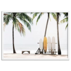 May include: Four surfboards leaning against a wooden rack on a sandy beach with two palm trees in the background. The surfboards are white and yellow.