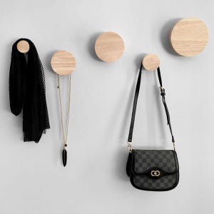 May include: Four wooden wall hooks with rounded edges, each with a different item hanging from them: a black scarf, a gold necklace, a black purse with a checkered pattern, and a black chain.