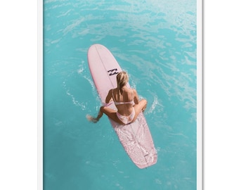 May include: A woman sits on a pink surfboard in the turquoise ocean. The surfboard has a white letter "N" on it.