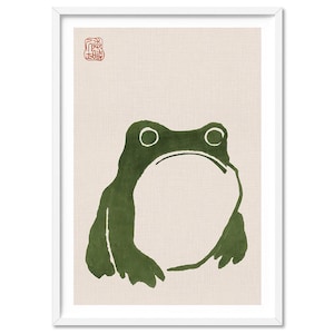 May include: A framed art print with a green frog illustration in a minimalist style. The frog is set against a beige backdrop. A red stamp-like symbol is in the upper left corner. The artwork is presented in a white frame.