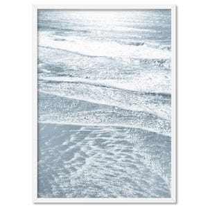 May include: A close-up photograph of a beach with blue-grey water and white foamy waves. The water is shallow and the waves are breaking gently on the shore.