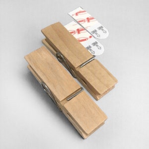 May include: Two wooden clothes pegs with metal springs. Each clothes peg has a white adhesive strip attached to it. The adhesive strips have the text "Command" and "3M" printed on them.