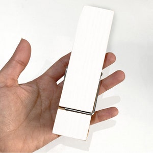 May include: White wooden clothes peg with a metal spring. The clothes peg is held in a hand.