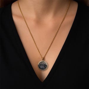 May include: A gold necklace with a circular pendant. The pendant has a detailed black and white design. The necklace is worn against a black top.