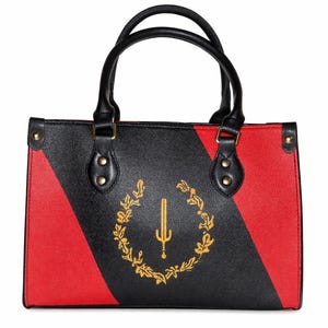 Puede incluir: Bolso de mano negro y rojo con asas negras. El bolso presenta un emblema dorado de una espada dentro de una corona. El diseño está dividido diagonalmente con una combinación de colores rojo y negro. El bolso tiene forma rectangular.