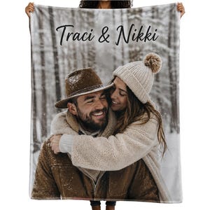 May include: A rectangular fleece blanket featuring a printed photograph of two people embracing in a snowy forest. The person on the left wears a brown hat and jacket, while the person on the right wears a beige beanie and coat. The text Traci & Nikki is printed in black script at the top.