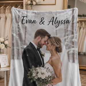 May include: A rectangular fleece blanket featuring a printed photograph of a couple in wedding attire standing in a snowy forest. The text Evan & Alyssa is printed in black script at the top. The couple is holding a bouquet of white roses.