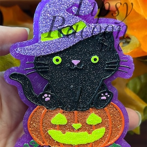 May include: A glittery purple and black hanging ornament featuring a black cat wearing a purple witch hat with a lime green band, sitting atop a glittery orange jack-o'-lantern with lime green eyes and mouth. The text Tipsy Raccoon is visible.