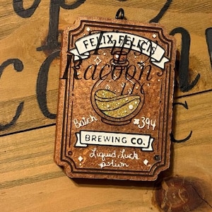 May include: A rectangular brown wooden ornament featuring a potion bottle illustration with gold glitter liquid. Text reads: FELIX FELICIS, Batch 394, BREWING CO., and Liquid luck potion. The design includes white banners and small star accents.