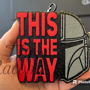 May include: A black and red keychain featuring the phrase "THIS IS THE WAY" in bold red letters. The right side of the keychain depicts a gray Mandalorian helmet. The keychain has a textured appearance and a metal ring.