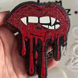 May include: A red glittery lips pendant with white teeth and dripping red blood. The pendant has a black outline and a small black loop at the top for hanging. The design is a spooky, Halloween-themed accessory.
