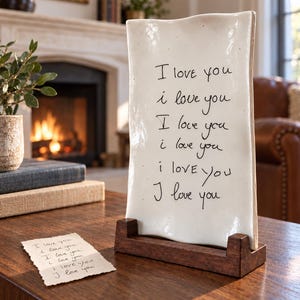 Op de afbeelding: Een rechthoekige witte keramische plaat met golvende randen, geplaatst op een donkerbruine houten standaard. De plaat bevat de handgeschreven zwarte tekst "I love you" die zes keer wordt herhaald. Een klein bijpassend briefje ligt plat in de buurt.